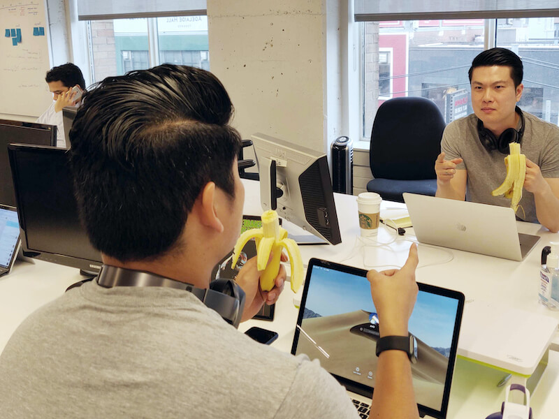 Two male team members in similar outfits and both holding a banana sit across from each other and point at each other